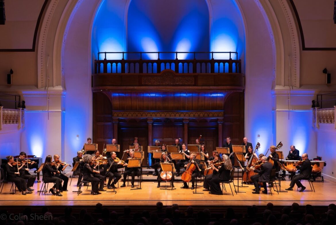 An orchestra sits on a stage with blue lighting behind