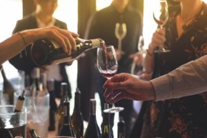 A hand pours a glass of red wine, while blurry figures stand holding wine glasses in the background