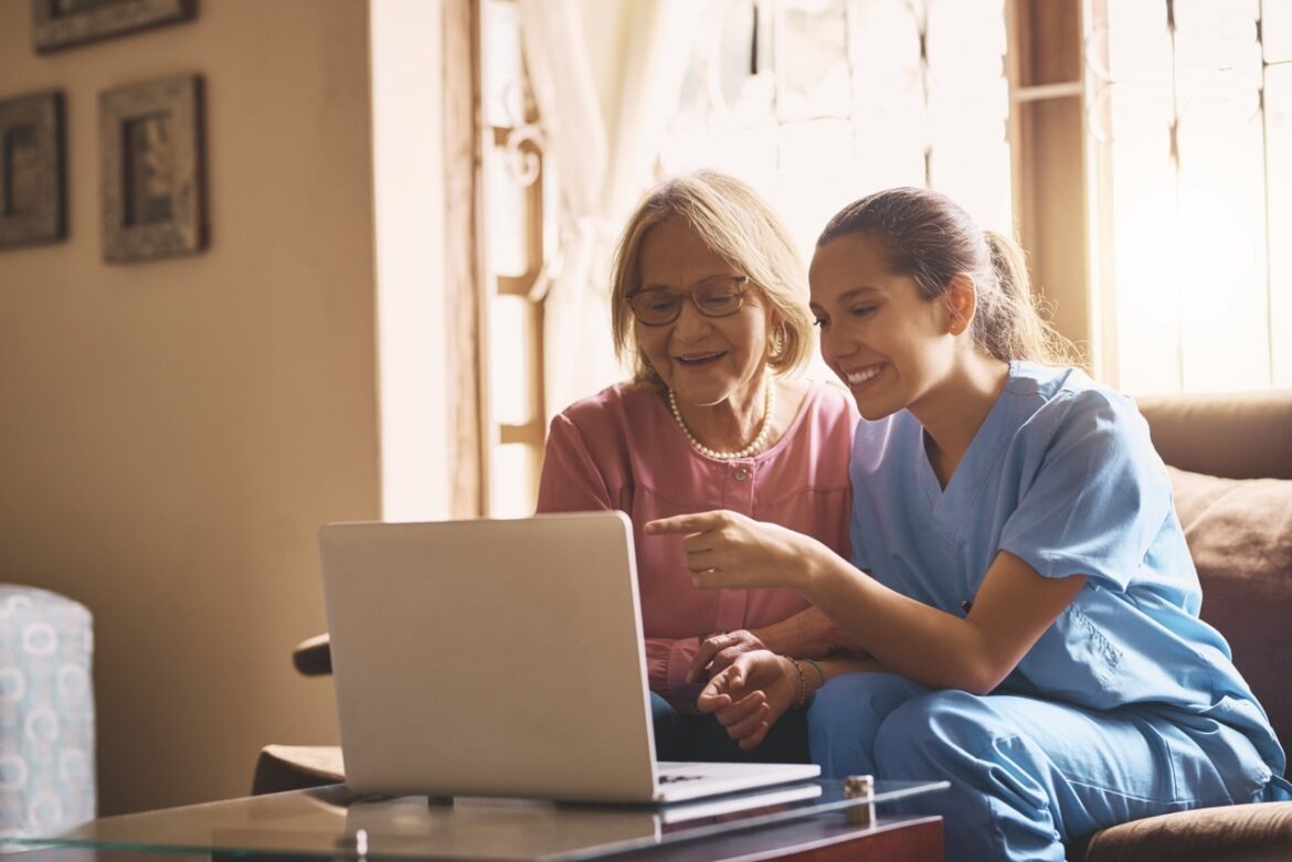 A nurse sits smiling with a senior woman. Both look at a laptop screen