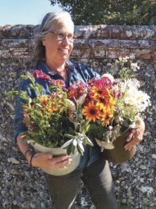 Portrait photo of a woman holding baskets of bright flowers in both hands, standing in front of a high stone wall