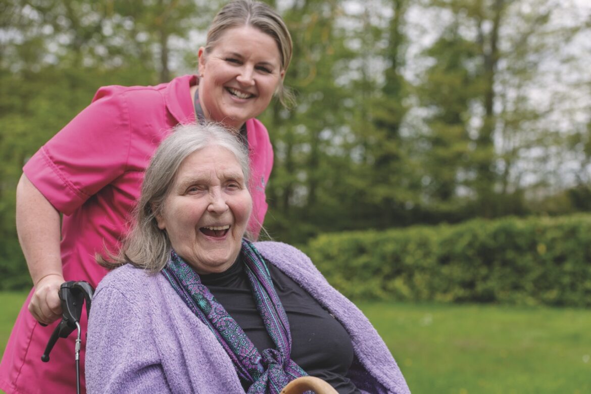 A senior lady wearing purple sitting in a wheelchair with a carer in pink standing behind smiling