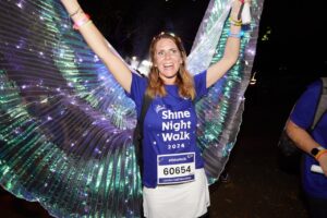 A woman wearing a purple t-shirt and glittery wings outside at night