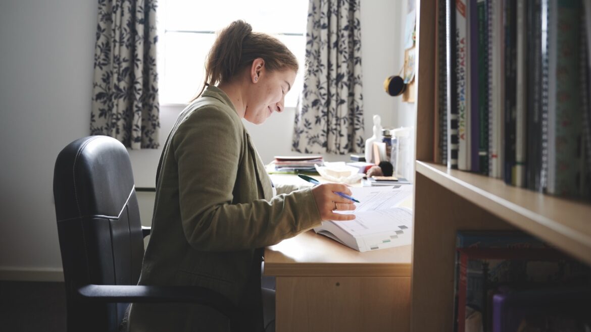 A woman sits at a desk wearing a green jacket and smiling as she flips through a textbook with a pen in her hand