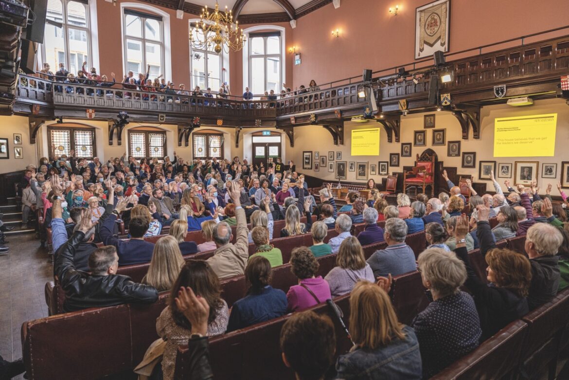 An ornate room with a gallery area on the second floor. The room is filled with a large seated crowd