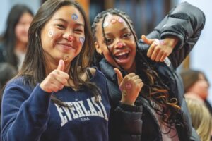 Two girls smile with their thumbs up. They have stickers decorating their face
