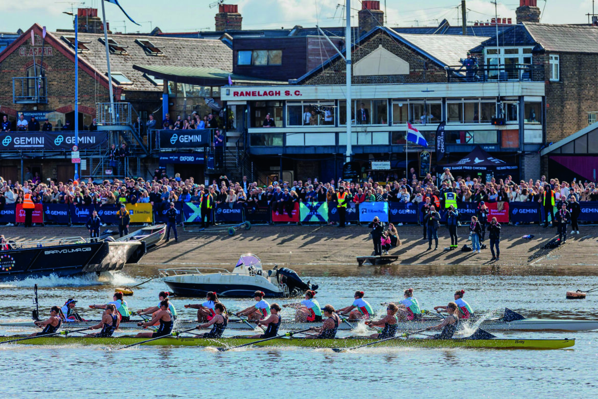 Two long rowing boats racing in a river. A large crowd stands on the bank watching
