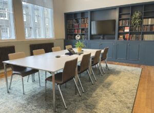 A modern office with a large conference table, a navy bookshelf and a TV screen