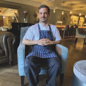 A photo of a man sitting with his hands clasped in a restaurant. He wears a white shirt and striped apron