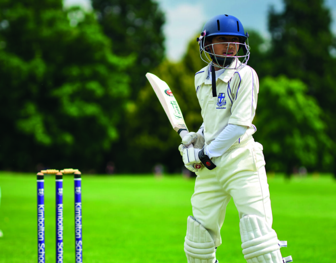 Two young boys playing cricket in a green field lined with trees