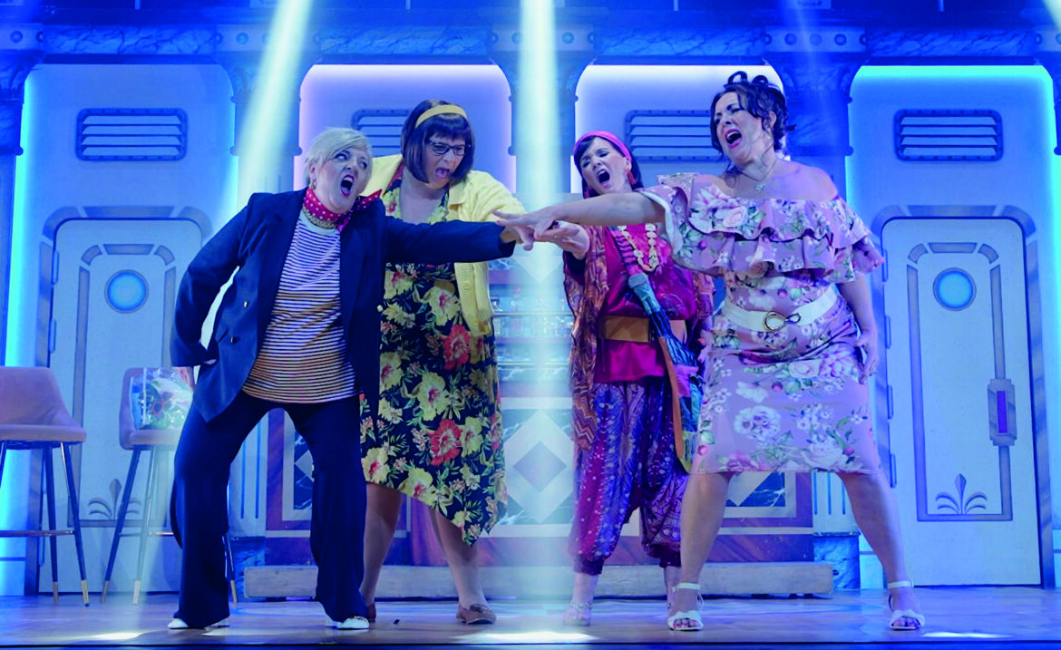 Four people stand on a stage in bright costumes. They reach their hands into the middle while singing