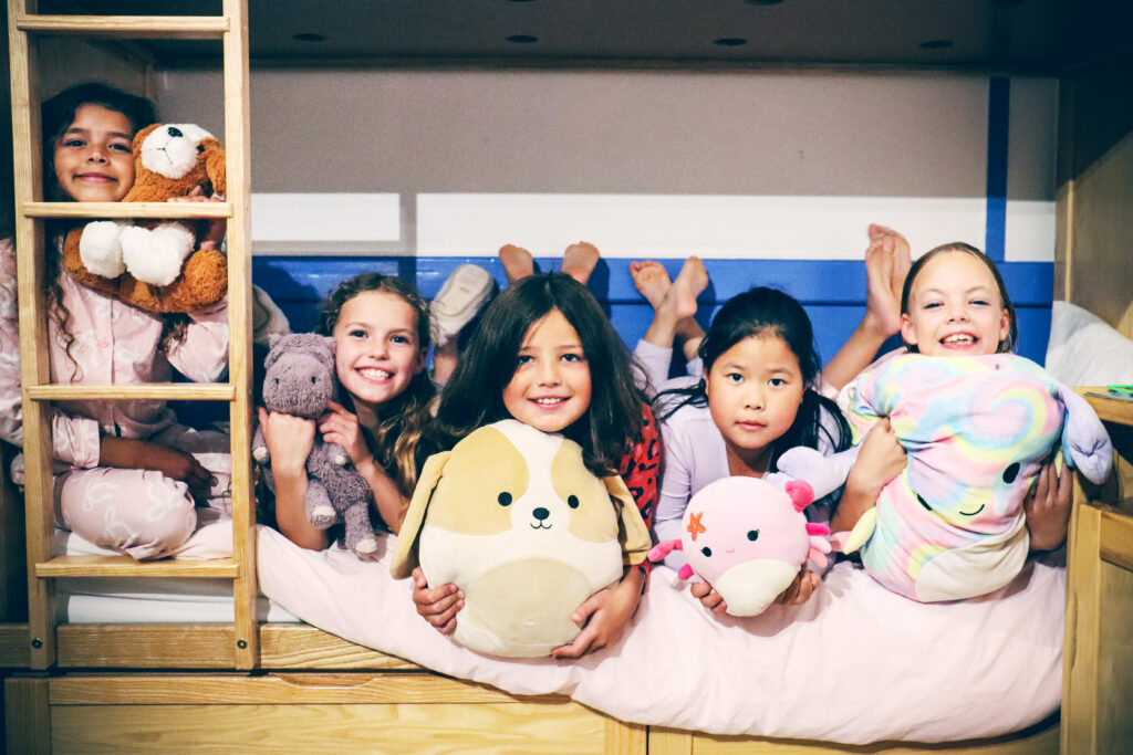 Five girls lay with stuffed animals on the lower level of a bunk bed. One girl pokes her head through the ladder