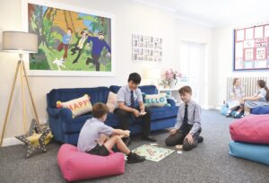 Three children in school uniform sit on bean bags, a sofa and the floor while they play Monopoly