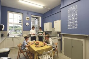 A group of children in a kitchen. Two boys sit at a table, while another two stand next to the sink with mugs