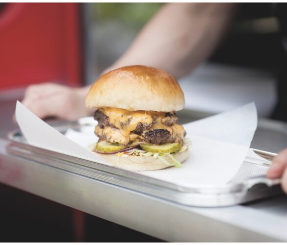 A burger on a napkin and metal tray. Two arms are either side