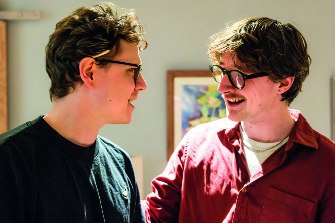 Two young men with glasses talk together while smiling. One has a cigarette behind his ear
