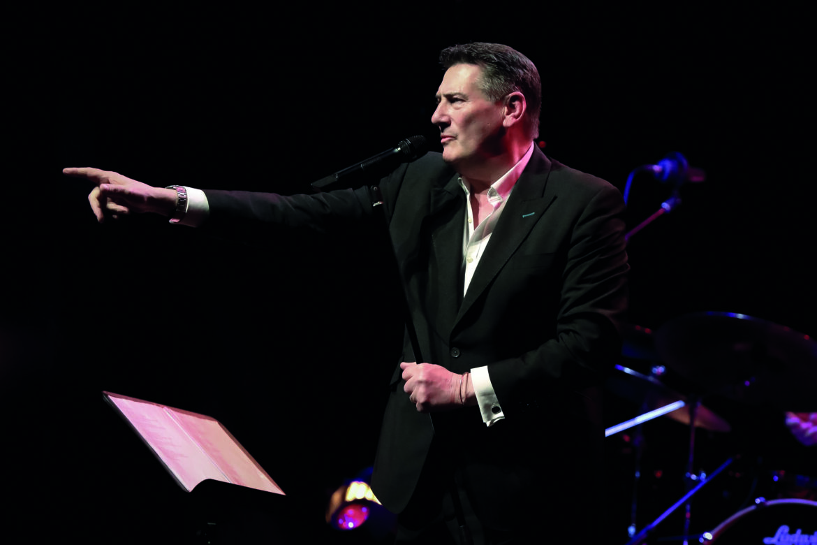 A man in a black suit pointing forward. He has a microphone and a sheet of music in front of him