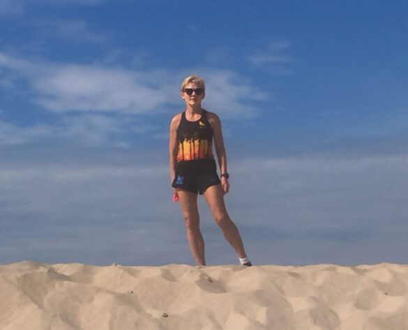 A woman standing on a beach, and wearing sports clothes and sunglasses