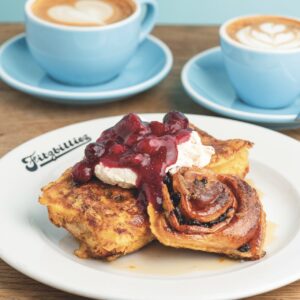 Chelsea buns sitting on a white plate. The buns are covered in jam and cream. Two blue mugs and saucers sit behind
