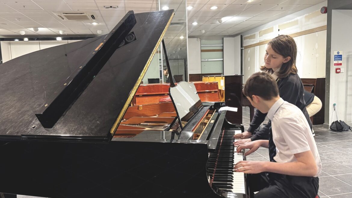 A boy sits on a stall playing a grand piano in a classroom while a girl stands next to him