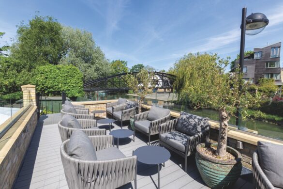 An outdoor seating area overlooking a river and bridge. The seating is made of grey armchairs and small round tables