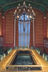 The inside of an ornate room with a square table with green carpets surrounding. A large window with red curtains is behind the table