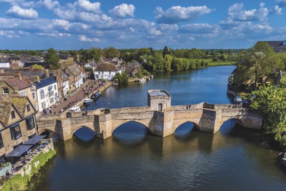 An arched bridge stretches over a river with houses on the left side and a wooded area on the right side
