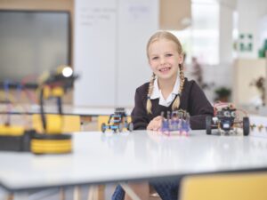 A smiling girl in school uniform sitting in a science lab with engineered cars in front of her