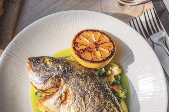 A top view of a dish showing fish on a bed of vegetables next to a grilled orange. A glass of wine and a bowl of olives sit on the table nearby
