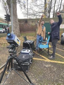 Two men stretch their arms, pointing upwards. A chair and camera are placed around them and they stand in a carpark on wet ground