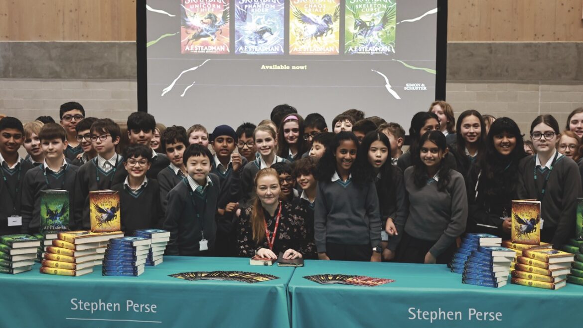 A group of students standing behind a blue table with a seated woman. A stack of books sit on the table