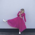 A woman in a bright pink dress kicking her leg out to the side. She holds a trumpet close to her heart