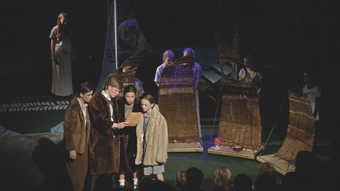 A group of children wearing long, furry coats standing onstage, looking at a letter. Children also stand in the background behind pieces of set