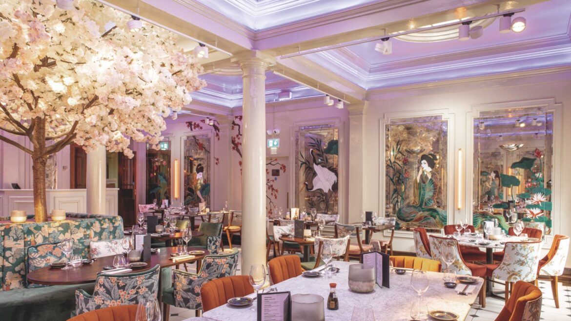 The interior of an expensive restaurant with artistic mirrors and flowery armchairs around tables. A large tree with white flowers is placed on the left side of the restaurant
