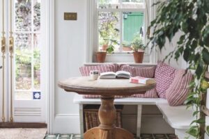 A bright kitchen area with a L-shaped bench with pink cushions and a round, wooden coffee table next to a glass door and window