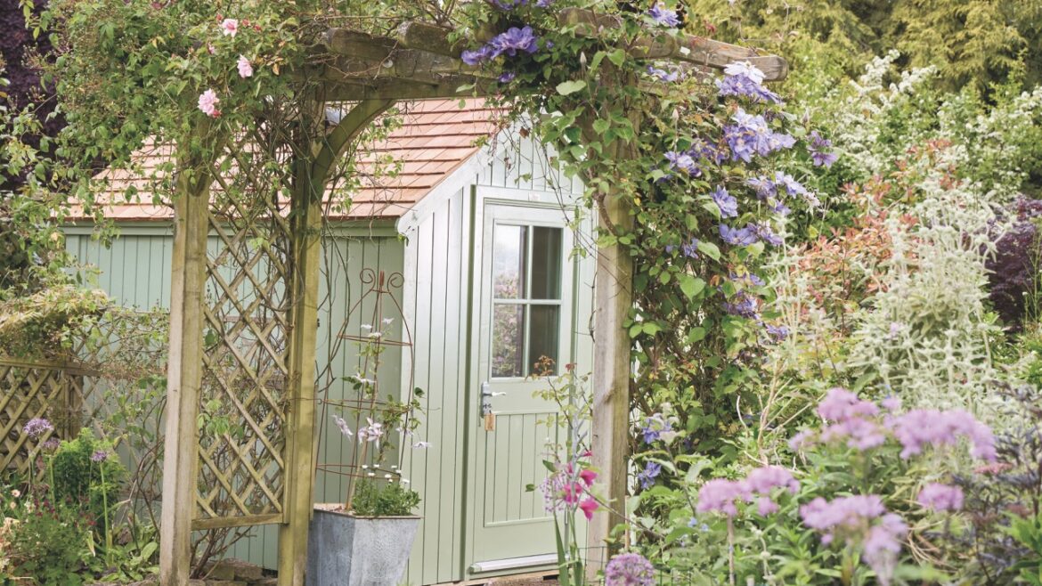 A light green shed in a flowery garden with a wooden arch leading further into the garden