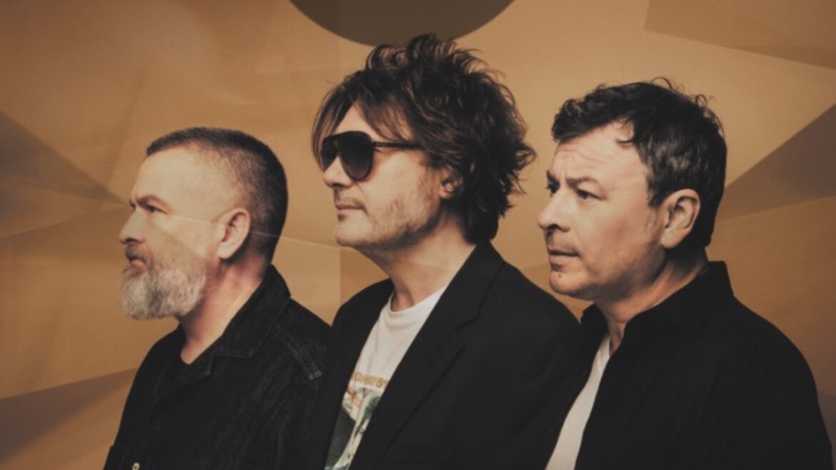 Three men stand in a row and look to the left. The man in the centre wears sunglasses. They stand in front of a textured brown background