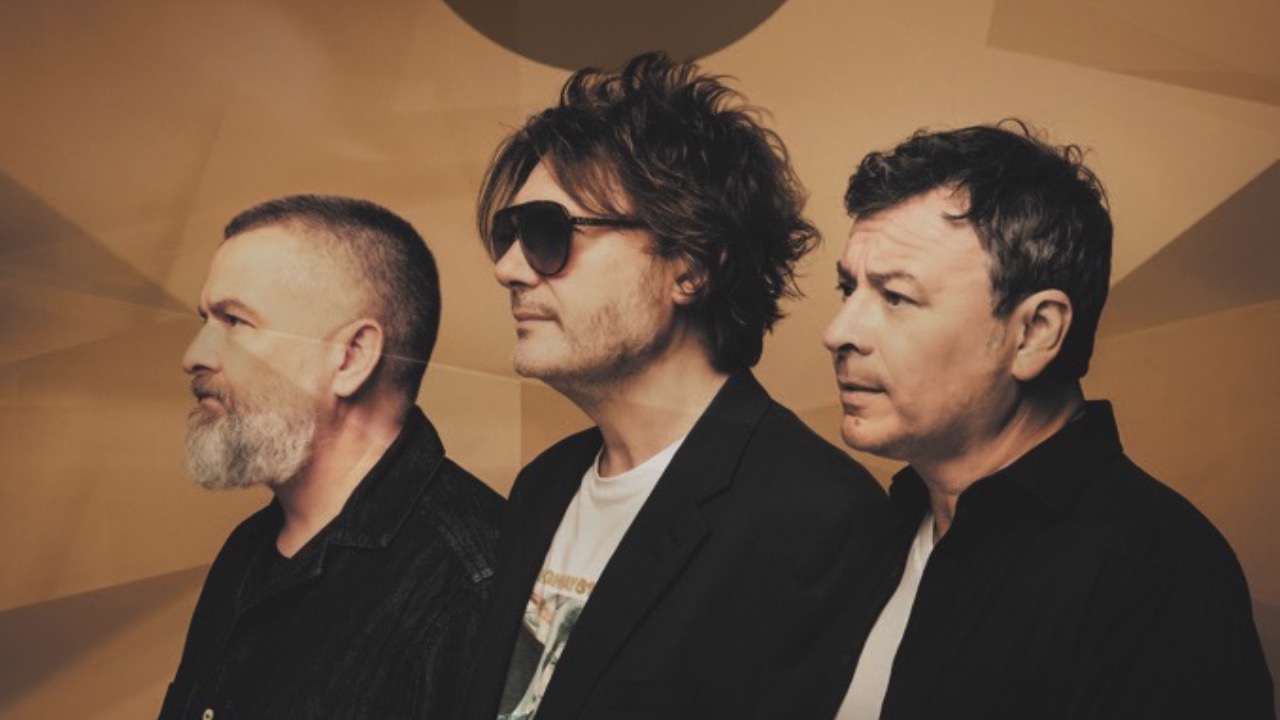 Three men stand in a row and look to the left. The man in the centre wears sunglasses. They stand in front of a textured brown background