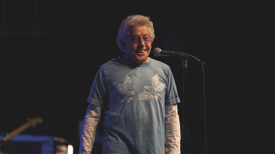 An old man with glasses wearing a blue T-shirt and a long undershirt walks towards a microphone on a dark stage