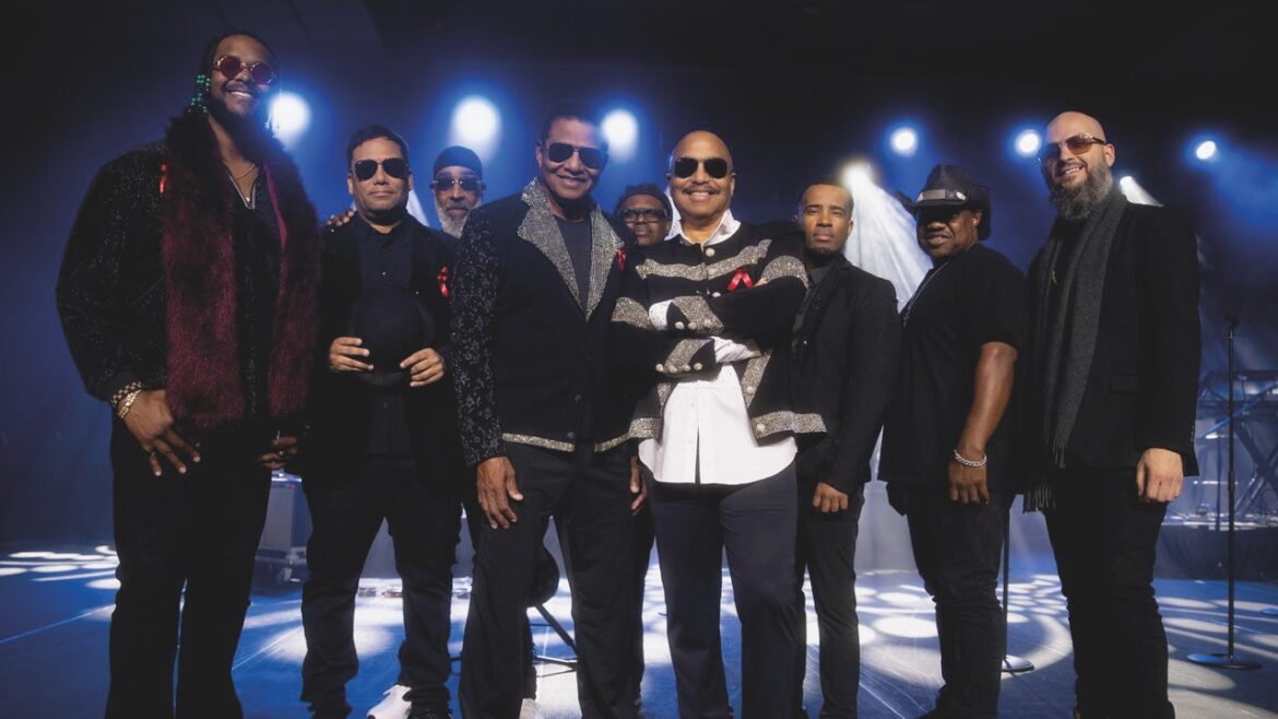 A group of men wearing stylish clothing a dark sunglasses stand as a group on a backlit stage