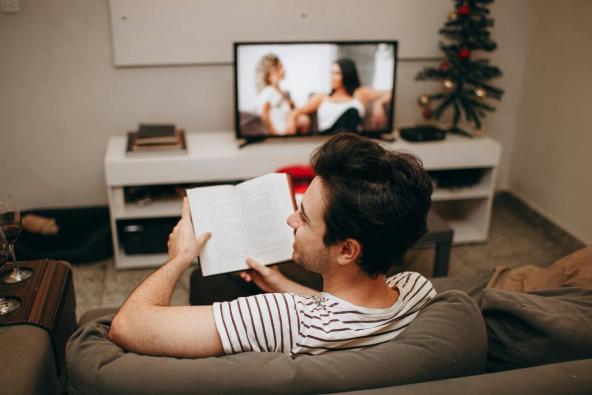 A man reading a book while watching a television in front of him in a living room
