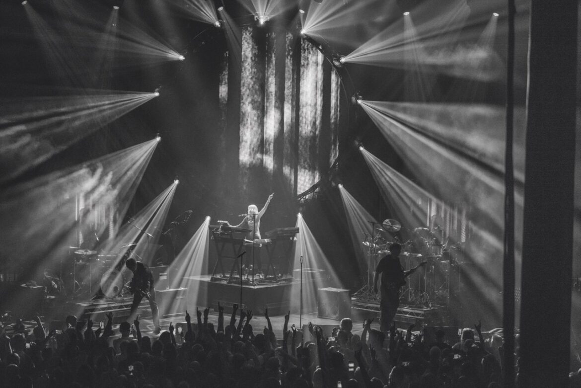 A black and white photo of the band Faithless standing with various instruments on a stage in front of a cheering crowd