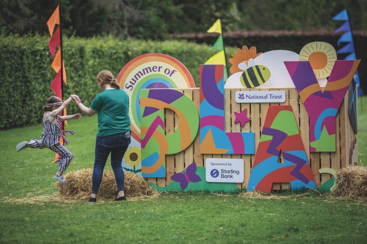 An adult helping a small child to jump in front of a wooden display by the National Trust in a field