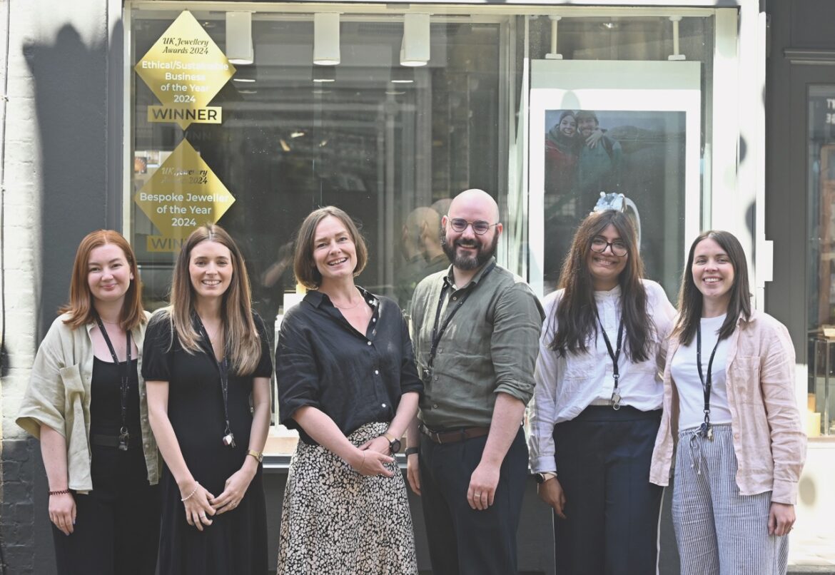 A group from Harriet Kelsall Bespoke Jewellery stands in a line smiling dress in work attire