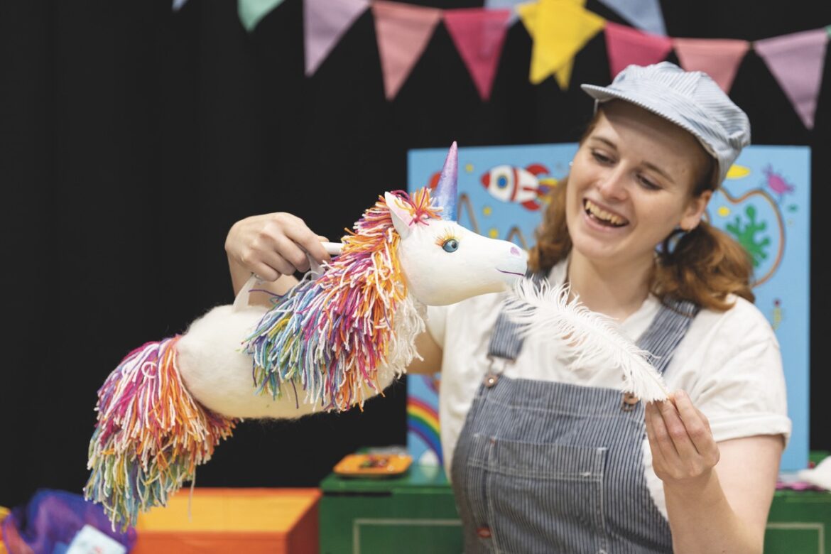 A woman in dungarees and a hat playing with a puppet unicorn and a feather as part of Pop-Up Adventures