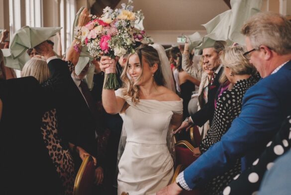 image1 Large A bride walks through a celebrating crowd in a large hall. She wears a short white wedding dress from The Pantry Bridalwear