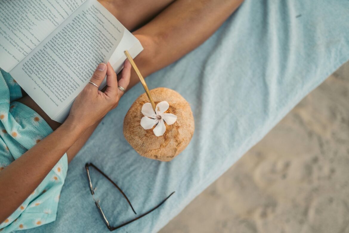 A top view of a person sitting on a towel on the beach reading a book. A pair of glasses and a coconut drink sits beside them