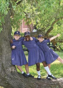Three young school girls in blue dresses and hats stand on a tree branch with their arms around each other 