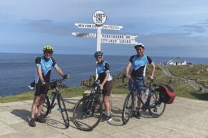 Three people standing next to a white destination signpost at the top of a cliff near the coast