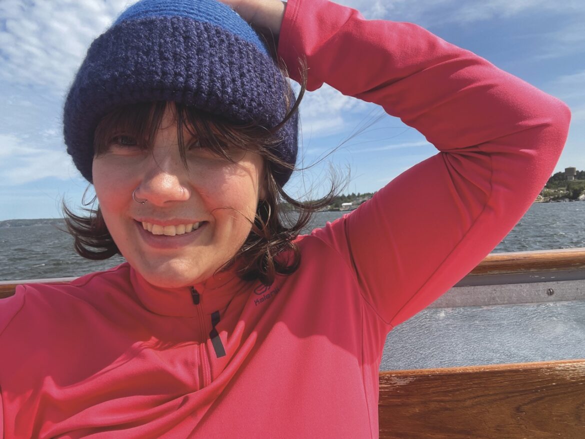 A woman standing in front of water, wearing a bright pink fleece and a blue hat. She holds her hat on as the wind tries to blow it away