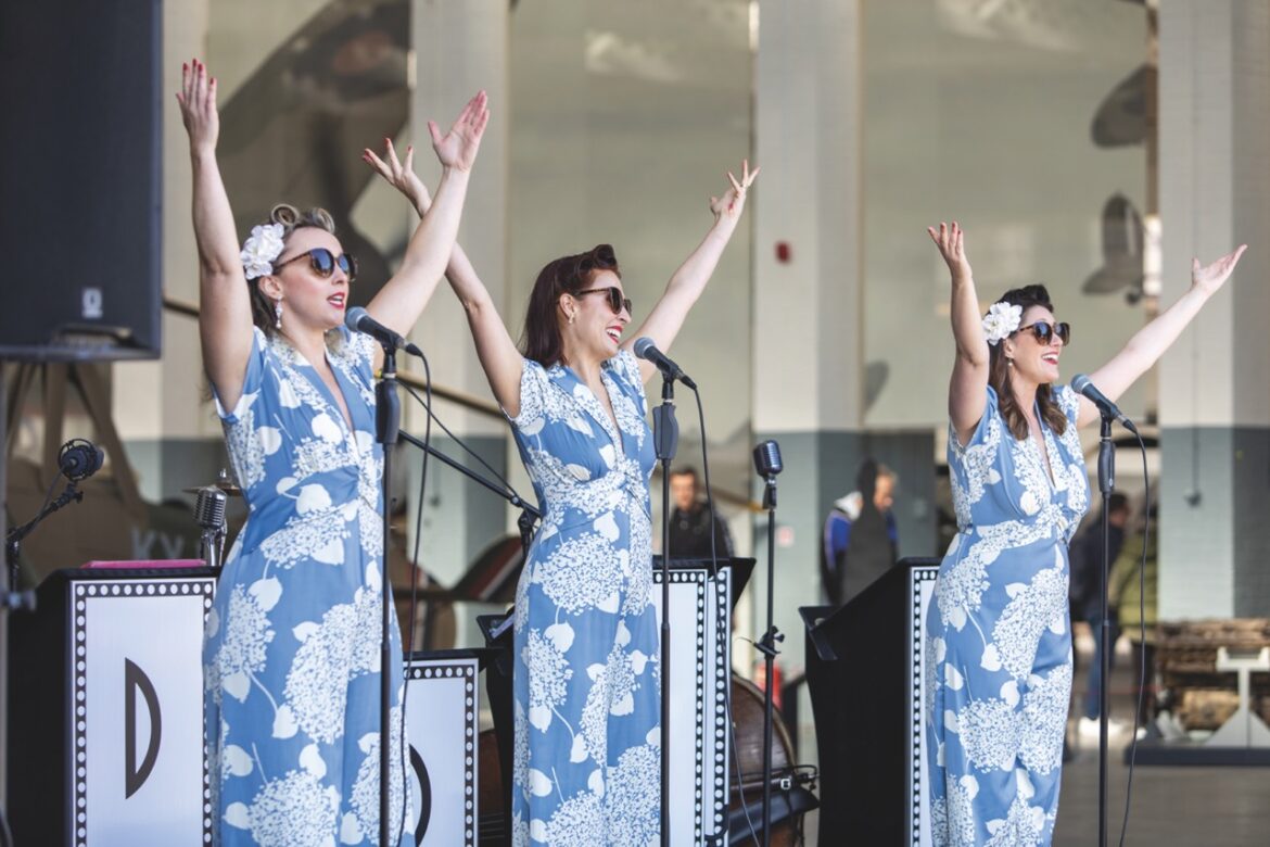 Three woman wearing blue and white jumpsuits, sunglasses and their hair in rollers. They sing on stage with their arms raised